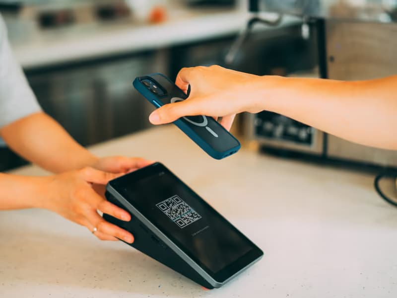 A hand holding a smartphone over a payment terminal showing a QR code