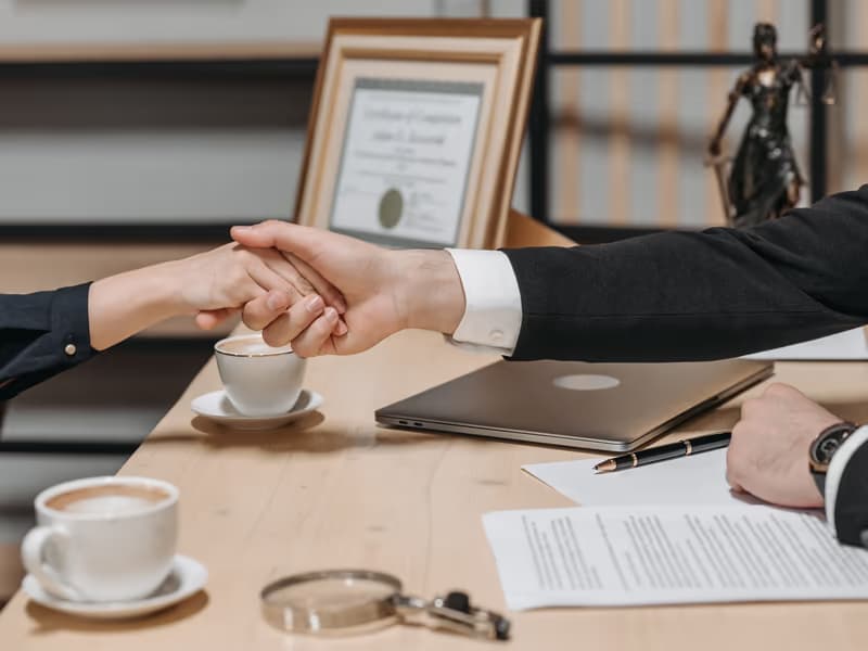 Handshake across a desk with scales of justice and a certificate behind